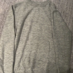 Grå stickad tröja med rund hals - En enkel och stilren grå stickad tröja med rund halsringning och långa ärmar. Tröjan har ribbade muddar vid ärmslut och nederkant, perfekt för lager-på-lager under kyliga dagar.