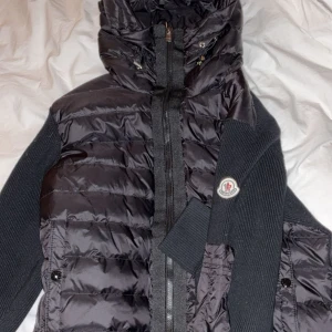 Svart Moncler pufferjacka med stickade ärmar - Svart pufferjacka från Moncler med stickade ärmar och sidor, quiltad framsida och huva. Jackan har dragkedja framtill, två fickor med knappar och Moncler-logga på ena ärmen. Perfekt för kyliga dagar med sin kombination av dun och stickat material. Kom med bud tack helt ny oanvänd