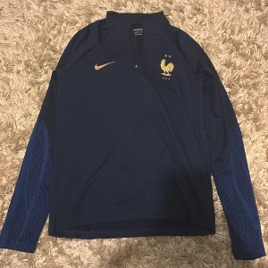 Frankrike Nike långärmad fotbollströja - Mörkblå långärmad fotbollströja från Nike med Frankrikes landslagsemblem och guldiga detaljer. Tröjan är i slim fit-modell och tillverkad i Dri-FIT-material som andas. Ärmarna har diskret mönster i blått och dragkedja vid halsen.