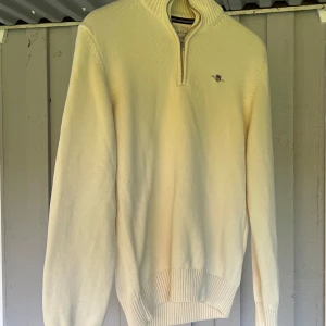 Ljusbeige stickad tröja från Gant - Säljer en ljusbeige stickad tröja från Gant med half zip och liten broderad logga på bröstet. Tröjan har hög krage, ribbade muddar och är långärmad. Perfekt för dig som gillar klassisk och clean stil.