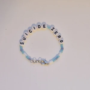 Pärlarmband Suicide Zero - Unikt pärlarmband med texten 'SUICIDE ZERO' i vita bokstavspärlor och små pärlor i blått och vitt. Armbandet har ett smidigt metallspänne och passar perfekt för dig som vill bära ett statement. Färgerna är fräscha och designen är enkel men snygg. Passar perfekt att ge bort till någon eller behålla själv. Storleken är ungefär 15-16,5 cm men är också gjorde med töjbar tråd.