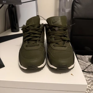 Nike Air Max 90 olivgröna sneakers - Säljer ett par Nike Air Max 90 sneakers i en snygg olivgrön färg med vita sulor. Skorna har klassisk Air Max-dämpning, snörning och är tillverkade i en mix av syntet och textil. Perfekta för dig som vill ha en stilren och bekväm sneaker med streetkänsla.