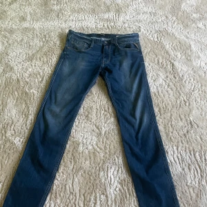 Blå Replay jeans Anbass herr 32/34 - Snygga blå jeans från Replay, modell Anbass. Klassisk femficksdesign med Replay-logga på bakfickan och läderpatch i midjan. Jeansen har en rak passform och är tillverkade i mjukt denimtyg för en bekväm känsla.