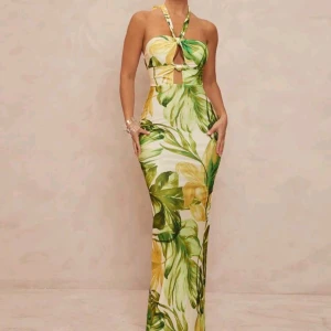 Grön och gul långklänning med snörning - Säljer en långklänning med tropiskt blommönster i grönt och gult på vit botten. Klänningen har halterneck, cutout framtill, snörning i ryggen och slits bak. Sitter tight och har en snygg passform, perfekt för sommarkvällar.