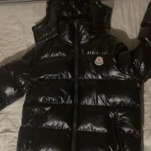 Svart Moncler pufferjacka med huva - Säljer en svart glansig pufferjacka från Moncler med huva och dragkedja framtill. Jackan har två fickor med dragkedja och klassisk Moncler-logga på bröstet. Perfekt för kalla dagar när du vill hålla stilen och värmen.