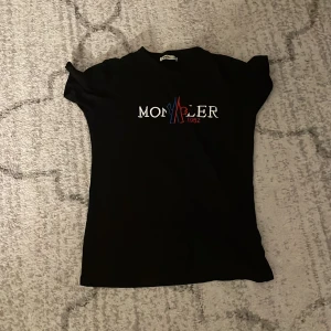 Svart Moncler t-shirt med tryck - Svart t-shirt från Moncler med klassiskt tryck i vitt, rött och blått framtill. T-shirten har rund halsringning och korta ärmar. Materialet känns mjukt och skönt, perfekt för en clean och stilren look.