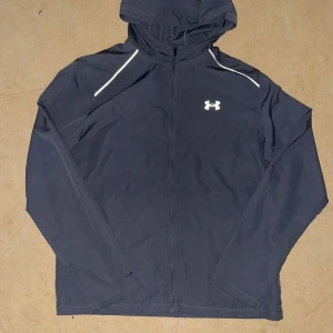 Svart vindjacka från Under Armour M - Säljer en svart vindjacka från Under Armour med huva och dragkedja framtill. Jackan har reflekterande detaljer vid axlarna, logga på bröstet och stort tryck på ryggen. Tillverkad i lätt och slitstark polyester, perfekt för aktiva dagar utomhus.