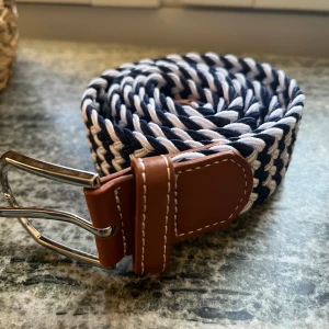 Randigt flätat bälte med skinn-detaljer - Snyggt flätat bälte i marinblått och vitt med bruna skinn-detaljer vid spännet. Bältet har ett coolt randigt mönster och ett silvrigt metallspänne. Perfekt accessoar för att lyfta din outfit med lite extra stil.