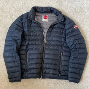 Mörkblå pufferjacka från Colmar | S/52 - Säljer en mörkblå pufferjacka från Colmar Originals. Jackan har quiltad design, två sidofickor med dragkedja och hög krage. Jackan har ett hål vid bakarmen, samt har ugglan lossnat!