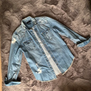 Ljusblå jeansskjorta med slitningar - Säljer en snygg ljusblå jeansskjorta från Zara Man med slitna detaljer och två bröstfickor med knappar. Skjortan har lång ärm och klassisk krage, perfekt för dig som gillar en cool och avslappnad stil.