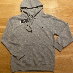 Grå hoodie från Polo Ralph Lauren - Klassisk grå hoodie från Polo Ralph Lauren med broderad logga på bröstet. Tröjan har huva med snörning, känguruficka och ribbade muddar. Tillverkad i mjuk bomullsmix som är perfekt för chill dagar.