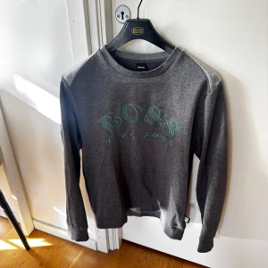Grå sweatshirt från Hugo Boss L - Snygg grå sweatshirt från Hugo Boss med grönt tryck på bröstet. Klassisk rund halsringning och långa ärmar. Perfekt för dig som gillar stilrena och bekväma plagg med premiumkänsla.