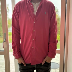 Rosa skjorta från Ralph Lauren - Säljer en snygg rosa skjorta från Ralph Lauren med klassisk krage och knappar framtill. Skjortan har lång ärm och ikonisk blå broderad logga på bröstet. Tillverkad i mjuk bomull som känns skön mot huden. Perfekt för dig som gillar stilrena och färgstarka plagg.