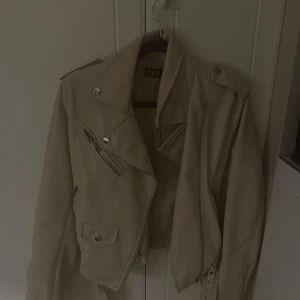 Beige skinnjacka från Zara - Snygg beige skinnjacka från Zara med coola detaljer som snedställda dragkedjor, knappar och axelklaffar. Jackan har klassisk bikerstil och är croppad i modellen. Perfekt för dig som vill ha en stilren och edgy look.