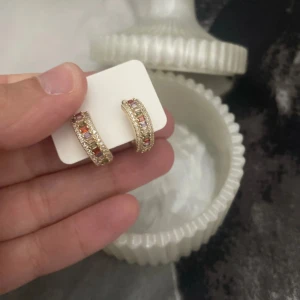 Guldiga örhängen med färgade stenar/ 18 k guldpläterade hoops - Snygga guldiga örhängen i halvbåge med små färgade stenar i rött, rosa och lila. Köpta i Turkiet! Örhängena har en glittrig look och detaljerade kanter som ger extra bling. Perfekta för dig som vill sticka ut med accessoarer som syns. 18 k guldpläterade 