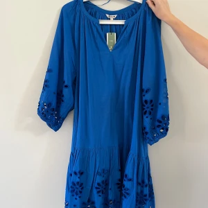 Blå klänning med broderade detaljer - Säljer en blå klänning med v-ringning och trekvartsärmar. Klänningen har broderade blommor och hålmönster på ärmar och nederdel, samt en luftig passform. Perfekt för dig som gillar färg och unika detaljer! Helt ny, lappen kvar. 