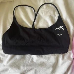 Svart sport-bh petch - Svart sport-bh med smala axelband som korsas i ryggen och en vit persikologga framtill. Enkel och stilren design, perfekt för stranden eller poolen. Materialet känns mjukt och följsamt mot huden.