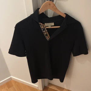 Svart Burberry pikétröja XS - Svart pikétröja från Burberry i klassisk modell med korta ärmar och krage. Ikoniskt rutigt tyg vid knäppningen och diskret broderad Burberry-logga på bröstet. Tillverkad i mjuk bomull för en skön känsla. Perfekt för en clean och stilren look. ÄKTA VAD JAG VET. 