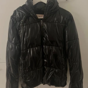 Svart pufferjacka från Nelly, stl S - Snygg svart pufferjacka från Nelly i glansigt material. Jackan har huva, dragkedja framtill och tryckknappar vid fickorna. de har lossnat färg från knapparna på fickorna. Det är lite ludd vid armvecket på armarna men det är inget som syns när man har på den. Det är även några defekter vid armarna och dragkedjan. Skriv till mig om du vill ha fler bilder kunde inte stoppa in alla