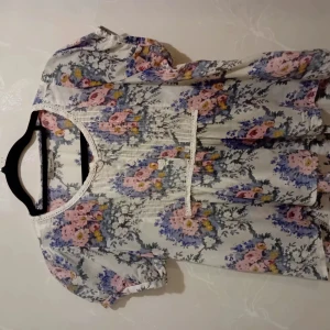 Blommig blus från N.Y. NearYou, stl 34 - Supersöt blommig blus i vitt med rosa, blå och gula blommor. Blusen har korta puffärmar, spetsdetaljer vid halsringning och framtill, samt små knappar. Materialet är tunn bomull, perfekt för vår och sommar. Söt och romantisk vibe!