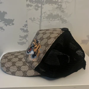 Gucci keps med tigertryck och mesh - Snygg keps från Gucci med klassiskt GG-mönster i beige och brunt, två tigrar framtill och svart mesh på sidorna och bak. Justerbar rem med grönt och rött band. Perfekt statement-piece för dig som vill sticka ut. Priset ej hugget! 