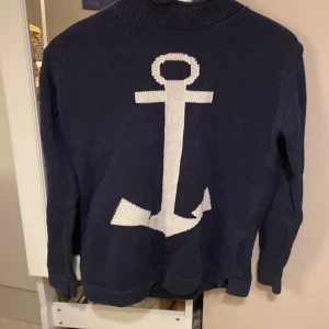 Marinblå stickad tröja med ankare - Säljer en marinblå stickad tröja från Pahladium i storlek M. Tröjan har ett stort vitt ankare på ryggen och är långärmad med ribbade muddar. Perfekt för dig som gillar nautisk stil och vill ha något snyggt och bekvämt.