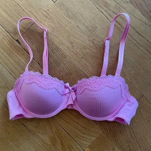 Rosa spets-bh från Hunkemöller 70A - Supersöt rosa bh från Hunkemöller med vadderade kupor, ribbat tyg och spetsdetaljer upptill. Dekorativ rosett framtill och justerbara axelband. Perfekt för dig som gillar feminina detaljer och pastellfärger. Toppen skick använd 1 gång.