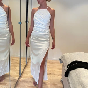 Vit långklänning med slits och snörning - Supersnygg vit långklänning i satinliknande material med smala axelband, öppen rygg med snörning och hög slits på ena sidan. Sitter figurnära och har en stilren, modern look som verkligen sticker ut.