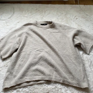 Beige stickad tröja med kort ärm - Säljer en beige stickad tröja från Weekday med korta ärmar och rund hals. Tröjan har en avslappnad passform och ribbade kanter vid hals, ärmslut och nederkant. Perfekt för lager-på-lager under hösten eller våren.