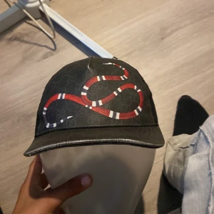 Gucci keps med orm och mesh, storlek L - Svart keps från Gucci med ikonisk röd och vit orm på framsidan. Kepsen har meshpaneler bak för extra ventilation och justerbart band med blå och röd rand. Märkesetikett på insidan. Perfekt statement-piece för dig som vill sticka ut.