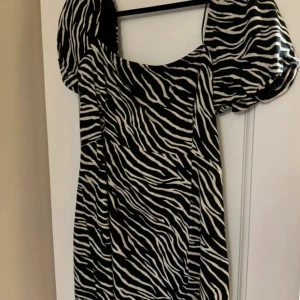 Zebramönstrad kortklänning från New yorker - Snygg kortklänning från New yorker med svartvitt zebramönster och puffiga korta ärmar. Klänningen har fyrkantig ringning, smockad rygg och dragkedja i sidan. Perfekt för dig som vill sticka ut med ett djurmönster. Storlek 40 men skulle säga att den är lite liten i storleken så mer en 38