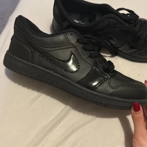 Nike Jordan 1 Low Triple Black - Säljer ett par helsvarta Nike Jordan 1 Low med klassisk siluett. Skorna har svart skinn, svarta snören och diskret Nike-logga på sidan. Perforerad tåbox för extra andningsförmåga och en robust yttersula med bra grepp. Perfekta för dig som gillar stilrena sneakers.