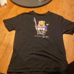 Svart Polo Bear t-shirt från Ralph Lauren - Svart t-shirt från Polo Ralph Lauren med ikoniska Polo Bear-trycket på bröstet. Björnen har skidutrustning och färgglada kläder. Klassisk rund hals och korta ärmar. Skön bomullskvalitet och avslappnad passform. Perfekt statement-plagg för dig som gillar streetstyle.