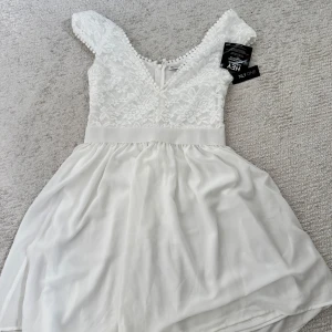 Vit spetsklänning från NLY One - Superfin vit kortklänning från NLY One med spets upptill, v-ringning och markerad midja. Kjolen är i lätt, flowigt tyg och klänningen har dekorativa bollar längs urringningen. Tillverkad i polyester och elastan för en bekväm passform.