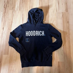 Hoodie från Hoodrich - Svart hoodie från Hoodrich med stor vit logga framtill och grafiskt tryck på ryggen. Tröjan har huva, känguruficka och ribbade muddar. Materialet är mjukt och skönt, perfekt för chill eller streetwear. Snygga blå och vita detaljer i trycket.