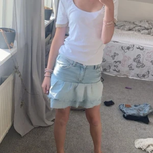 Ljusblå jeanskjol med volang - Supersöt ljusblå jeanskjol med volangkant och råa kanter. Kjolen har klassiska jeansdetaljer som fickor och bälteshällor. Perfekt för dig som gillar en avslappnad och trendig stil.