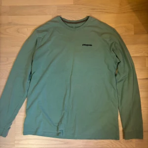Grön långärmad tröja från Patagonia - Snygg grön långärmad tröja från Patagonia med klassisk logga på bröstet och stort tryck på ryggen. Tröjan är i bomull och har en relaxed passform, perfekt för chill dagar. Rund halsringning och mjukt material gör den superbekväm.