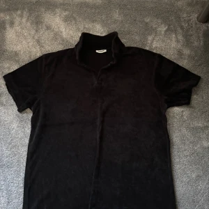 Svart frotté t-shirt från KappAhl - Svart t-shirt från KappAhl i mjuk frotté med korta ärmar och krage. Skön och avslappnad passform, perfekt för chill dagar. Enkel design utan mönster eller tryck, passar dig som gillar stilren look. Skicket är 9,5/10 och använd 2 gånger. 