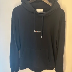 Svart hoodie från Saint Laurent storlek L - Svart hoodie från Saint Laurent med broderad logga på bröstet. Klassisk känguruficka framtill, justerbar huva med snörning och långa ärmar. Tillverkad i mjuk bomull för en skön och avslappnad look.