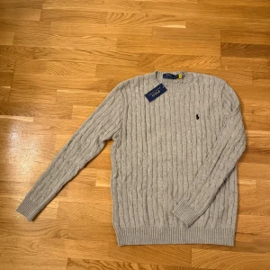 Grå stickad tröja Polo Ralph Lauren - Kabelstickad grå tröja från Polo Ralph Lauren med rund halsringning och broderad logga på bröstet. Tröjan är långärmad och har ribbade muddar vid ärmslut och nederkant. Perfekt för lager-på-lager och snygg till jeans.