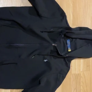 Svart hoodie från Polo Ralph Lauren - Svart hoodie från Polo Ralph Lauren med dragkedja och huva. Klassisk design med broderad logga på bröstet och justerbara snören i huvan. Perfekt för en avslappnad och stilren look.