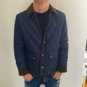 Quiltad blå jacka Polo Ralph Lauren - Snygg quiltad jacka från Polo Ralph Lauren i mörkblått med bruna detaljer och krage i manchester. Jackan har tryckknappar framtill, två stora fickor och klassisk röd logga på bröstet. Perfekt för höst och vår när du vill ha en stilren look. Modellen är runt 168 cm och väger 53 kg det är bara att höra av sig vid frågor och funderingar 😀