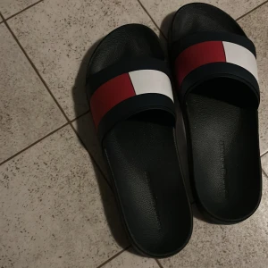 Tommy Hilfiger slides i rött, vitt, blått - Snygga slides från Tommy Hilfiger med bred rem i rött, vitt och mörkblått. Skorna har svart sula med mönstrad undersida och klassisk logga på remmen. Perfekta för sommaren eller att chilla i hemma. 