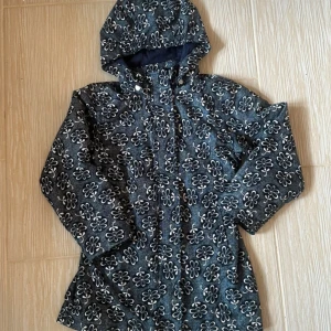 Blommig jacka med huva från Name It - Mörkblå jacka från Name It med vita blommor över hela tyget. Jackan har huva, dragkedja framtill och är tillverkad i polyester med bomullsfoder. Perfekt för kyliga dagar och har en snygg, lekfull look.