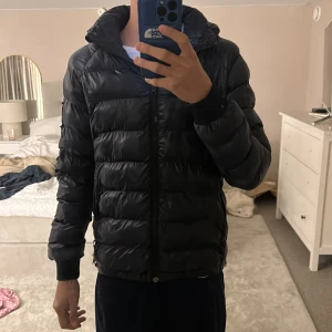 Blå pufferjacka från Peak Performance - Säljer en blå pufferjacka från Peak Performance med huva och dragkedja. Jackan är vadderad och har en glansig finish, perfekt för kyliga dagar. Klassisk design med diskreta logodetaljer och ribbade muddar vid ärmsluten. Ett litet märka på jackan som kan fixas. Storlek 160 men passar 165-170.