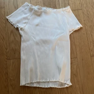 Vit ribbad offshoulder topp Lindex - Säljer en vit ribbad offshoulder topp från Lindex i storlek 146/152. Toppen har korta ärmar och en lätt vågig kant vid halsringning, ärmslut och nederkant. Perfekt för dig som gillar en enkel men trendig stil.