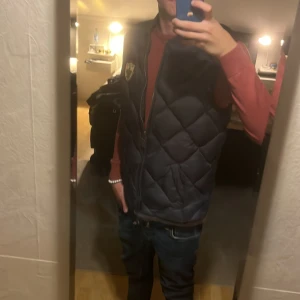 Mörkblå dunväst från Morris Tradition - Säljer en snygg mörkblå dunväst från Morris Tradition med quiltad design och brunt kantband. Västen har ett broderat Morris-emblem på bröstet och dragkedja framtill. Perfekt för lager-på-lager och stilren look. Storlek medium sitter som Small!