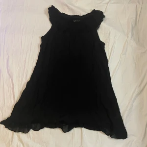 Svart ärmlös klänning  - Svart, luftig och ärmlös klänning med volangdetaljer vid halsringning och fåll. A-linjeformad siluett som ger en avslappnad och trendig look. Perfekt för dig som gillar stilrena och enkla plagg med en twist.