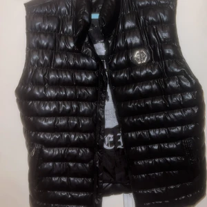 Svart dunväst från Philipp Plein - Snygg svart dunväst från Philipp Plein med glansig finish och quiltad design. Västen har dragkedja, två fickor och ett stort döskalletryck på insidan. Märkets logga syns både fram och bak. Perfekt för dig som vill sticka ut med en exklusiv streetstyle-look.