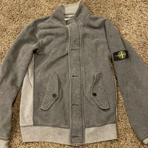 Grå sweatshirtjacka från Stone Island - Grå sweatshirtjacka från Stone Island med klassisk patch på ärmen. Jackan har hög krage, knäppning framtill och två fickor med lock och knapp. Mjuk bomullskänsla och ribbade muddar vid ärmslut och nederkant. Perfekt för dig som gillar streetwear.
